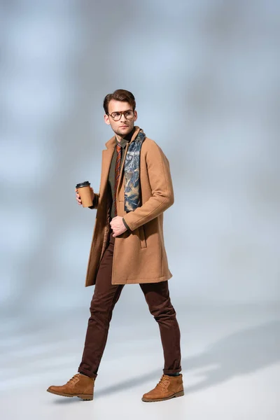 Stylish man in winter coat holding coffee to go and walking on grey — Stock Photo