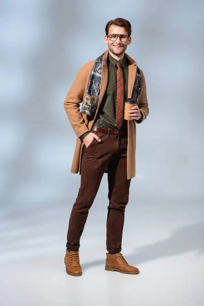 Полный рост счастливого человека в зимнем пальто, держащего бумажный стаканчик, стоя с рукой в кармане на сером — стоковое фото