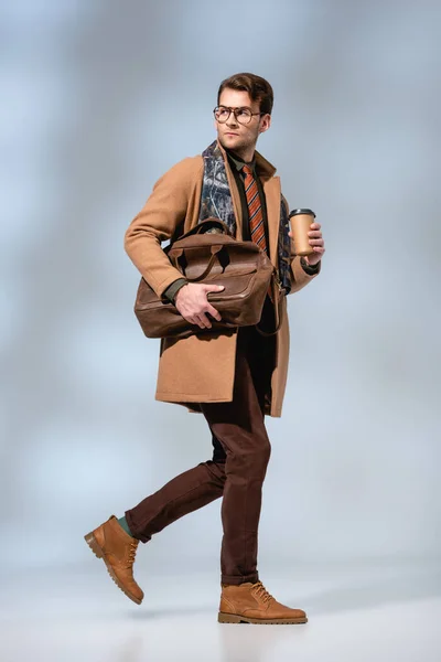 Longueur totale de l'homme élégant en manteau d'hiver tenant tasse en papier et serviette en cuir tout en marchant sur gris — Photo de stock