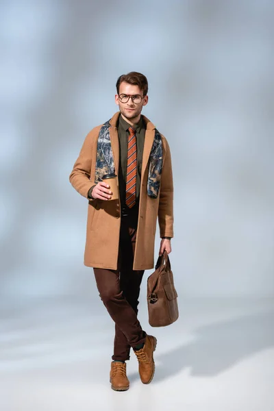 Full length of stylish man in winter coat holding paper cup and leather briefcase while standing with crossed legs on grey — Stock Photo