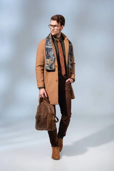 Повна довжина стильного чоловіка в зимовому пальто тримає шкіряний портфель і стоїть з рукою в кишені на сірому — стокове фото