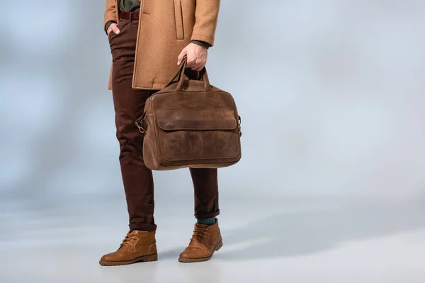 Частичный вид стильного мужчины, держащего кожаный портфель и стоящего с рукой в кармане на сером — стоковое фото