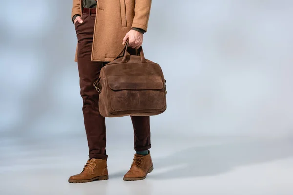 Vista parcial del hombre elegante que sostiene el maletín de cuero y de pie con la mano en el bolsillo en gris - foto de stock