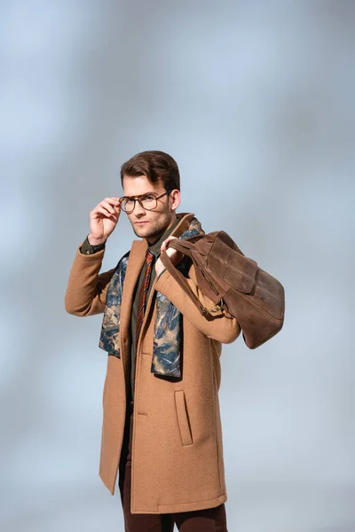 Stylish man in winter coat adjusting glasses while holding leather briefcase on grey — Stock Photo