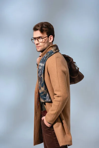 Стильный мужчина в зимнем пальто и очки, стоящие с рукой в кармане держа кожаный портфель на сером — стоковое фото