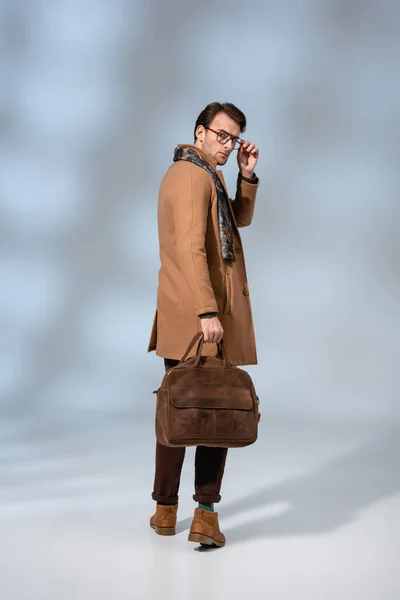 Полная длина стильный мужчина в зимнем пальто регулировки очки, держа кожаный портфель на сером — стоковое фото