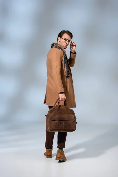 Toute la longueur de l'homme élégant dans des lunettes de réglage de manteau d'hiver tout en tenant la mallette en cuir sur gris — Photo de stock