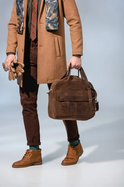 Обрізаний вид стильного чоловіка в зимовому пальто, що тримає шкіряний портфель і рукавички на сірому — стокове фото