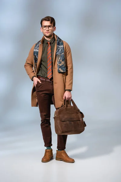 Full length of stylish man in winter coat holding leather briefcase and gloves while posing with hand in pocket on grey — Stock Photo