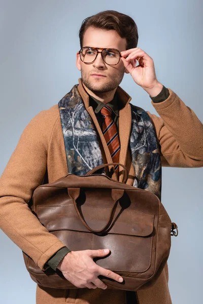 Hombre elegante en abrigo de invierno ajuste de gafas mientras sostiene el bolso marrón en gris - foto de stock