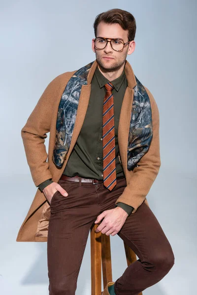 Élégant homme en manteau d'hiver et lunettes assis sur une chaise en bois avec la main dans la poche sur gris — Photo de stock