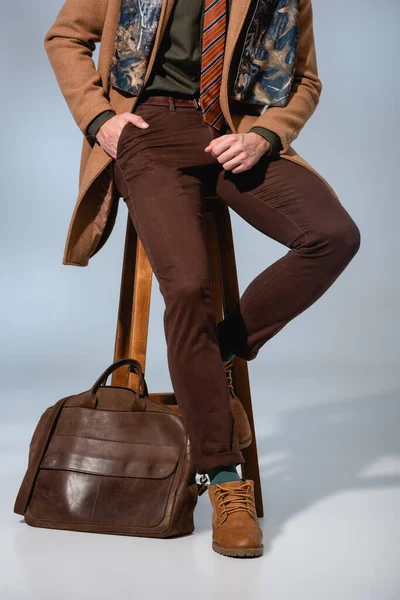 Cropped view of stylish man in winter coat sitting on wooden chair with hand in pocket near briefcase on grey — Stock Photo