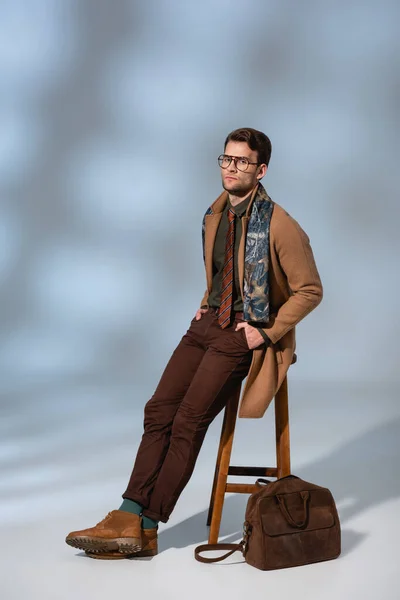 Full length of trendy man in winter coat and glasses sitting on wooden chair with hands in pockets near briefcase on grey — Stock Photo