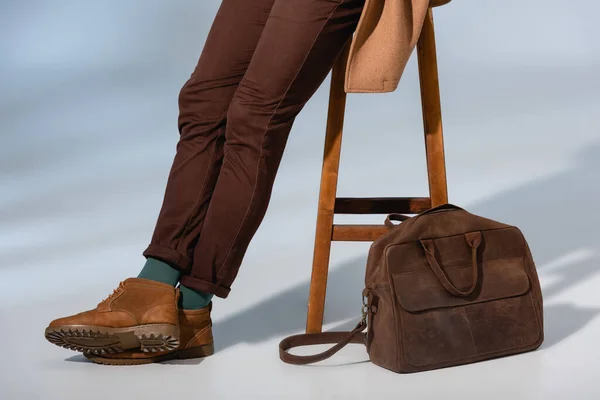 Vue recadrée de l'homme élégant en manteau d'hiver assis sur une chaise en bois près de mallette sur gris — Photo de stock