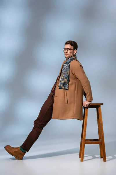 Full length of trendy man in winter coat and glasses leaning on wooden chair on grey — Stock Photo