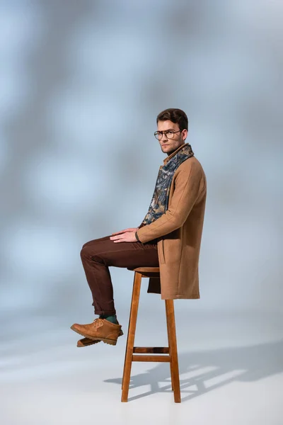 Toute la longueur de l'homme élégant en tenue d'hiver et des lunettes assis sur une chaise en bois sur gris — Photo de stock