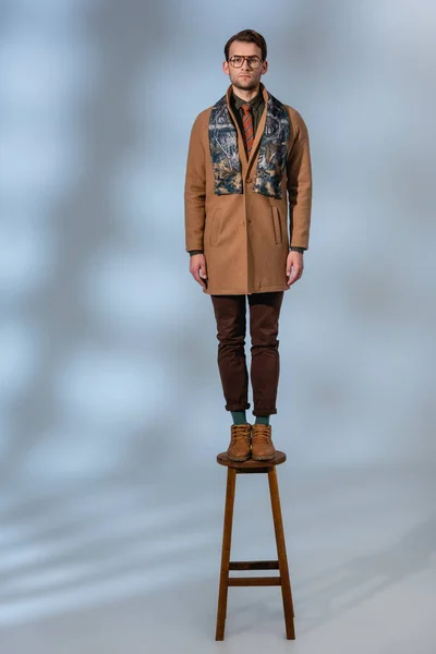Comprimento total do homem elegante em roupas de inverno e óculos de pé em banquinho de madeira em cinza — Fotografia de Stock