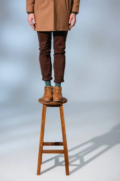 Обрізаний вид стильного чоловіка в зимовому вбранні, що стоїть на дерев'яному табуреті на сірому — стокове фото
