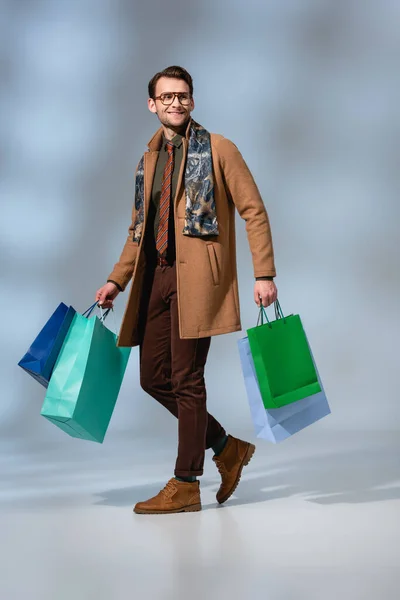 Full length of cheerful customer in glasses holding paper bags on grey — Stock Photo