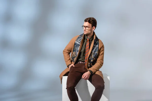 Стильный мужчина в очках и зимнем пальто сидит с рукой в кармане на белом кубе на сером фоне — стоковое фото