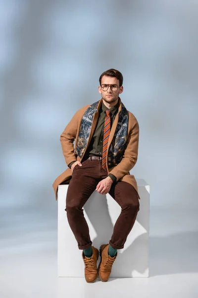 Homme élégant dans des lunettes et manteau d'hiver assis avec la main dans la poche sur cube blanc sur gris — Photo de stock