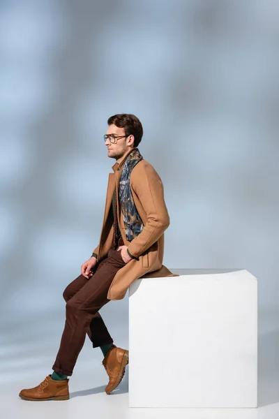 Hombre de moda en gafas y abrigo de invierno sentado con la mano en el bolsillo en cubo blanco en gris - foto de stock