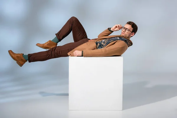 Hombre con estilo en gafas y abrigo de invierno acostado en cubo blanco sobre fondo gris - foto de stock