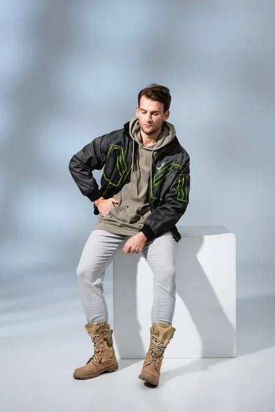 Full length of trendy man in hoodie and parka with hand in pocket sitting on white cube on grey — Stock Photo