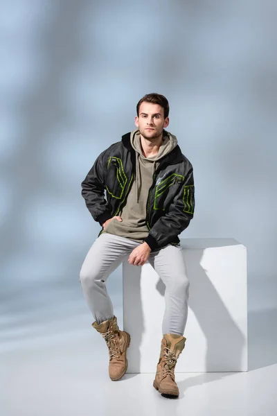 Longitud completa del hombre de moda en botas, sudadera con capucha y parka posando mientras está sentado en cubo blanco en gris - foto de stock