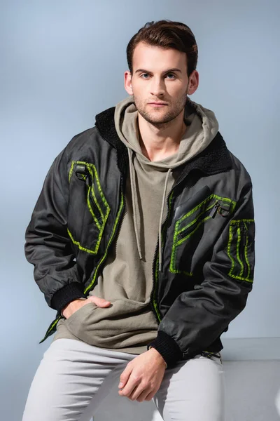 Trendy man in hoodie and parka posing while sitting on white cube isolated on grey — Stock Photo