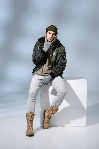 Longitud completa del hombre de moda en sombrero, guantes y parka posando mientras está sentado en cubo blanco en gris - foto de stock