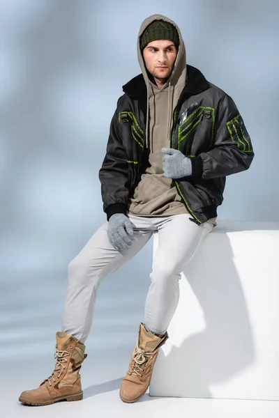 Longitud completa del hombre de moda en sombrero, guantes y parka sentado en cubo blanco en gris - foto de stock