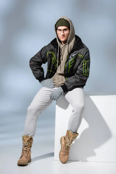 Comprimento total do homem na moda em chapéu, luvas e parka sentado com a mão no quadril no cubo branco no cinza — Fotografia de Stock