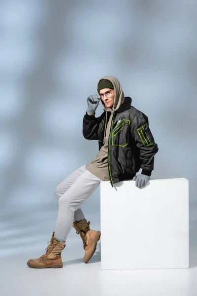 Full length of trendy man in hat, gloves and parka adjusting glasses while sitting on white cube on grey — Stock Photo