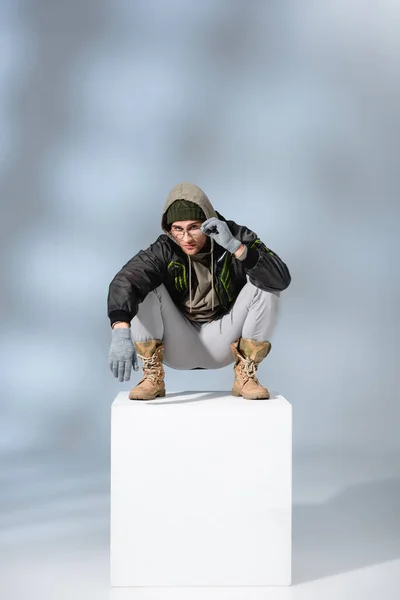 Stylish man in hat, gloves and parka adjusting glasses while sitting on white cube on grey — Stock Photo