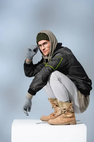 Stylish man in hat, gloves and anorak adjusting glasses while sitting on white cube on grey — Stock Photo