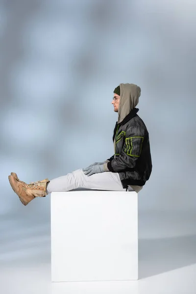 Side view of trendy man in hat, gloves and anorak sitting on white cube on grey — Stock Photo