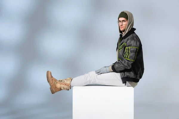 Young man in hat, gloves and anorak sitting on white cube on grey — Stock Photo