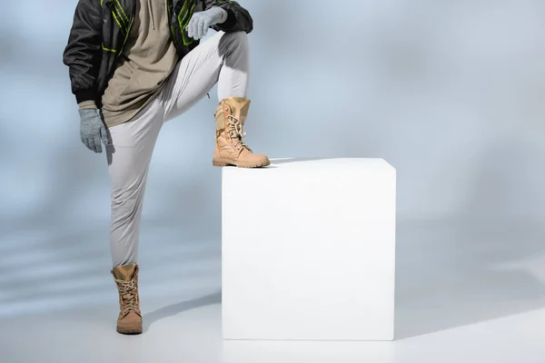 Vista recortada de hombre joven de moda en sombrero, guantes y anorak apoyado en cubo blanco sobre gris - foto de stock