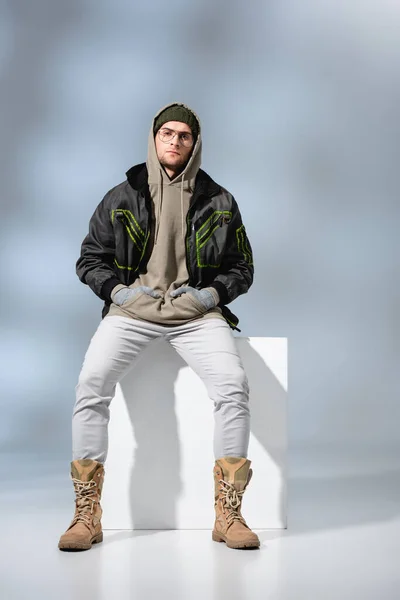 Piena lunghezza di giovane uomo di tendenza in cappello e anorak seduto con le mani in tasche sul cubo bianco su grigio — Foto stock