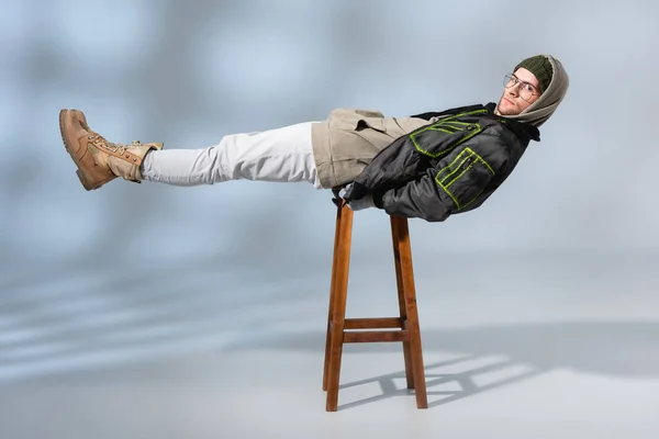 Young trendy man in hat and anorak lying horizontally on wooden stool on grey — Stock Photo