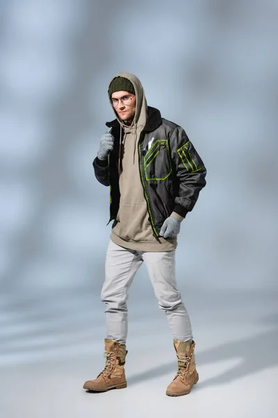 Full length of young trendy man in hat, hoodie and anorak standing on grey — Stock Photo