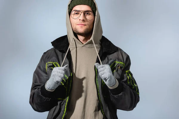 Homme à la mode dans des lunettes, chapeau, anorak et gants regardant la caméra sur gris — Photo de stock