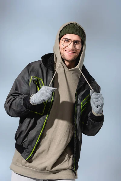Модный человек в очках, шляпе, анораке и перчатках, улыбающийся на сером — стоковое фото