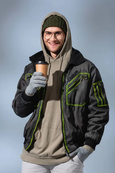 Happy man in glasses, hat, anorak and gloves holding paper cup while standing with hand in pocket on grey — Stock Photo