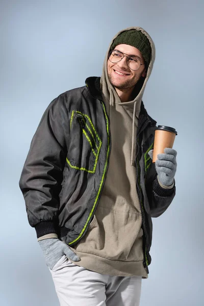 Веселый человек в очках, шляпе, анораке и перчатках держа бумажную чашку, стоя с рукой в кармане на сером — стоковое фото