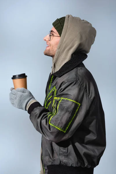 Side view of happy man in glasses, hat, anorak and gloves holding paper cup isolated on grey — Stock Photo