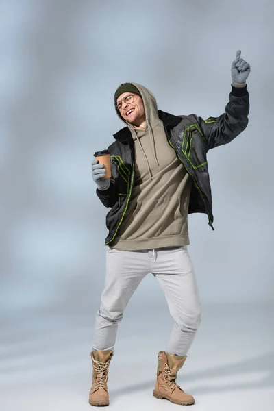 Comprimento total de jovem feliz homem de chapéu, capuz e anorak segurando copo de papel e dançando em cinza — Fotografia de Stock