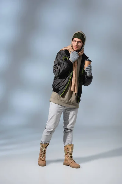 Full length of young man in hat, hoodie and anorak standing and holding coffee to go on grey — Stock Photo