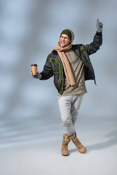 Pleine longueur de l'homme excité en chapeau, sweat à capuche et anorak tenant tasse en papier sur gris — Photo de stock